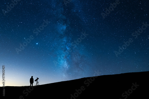 Person observing the blue starry sky with a telescope at night. Silhouette of an astronomer observing the immensity of the universe and the Milky Way