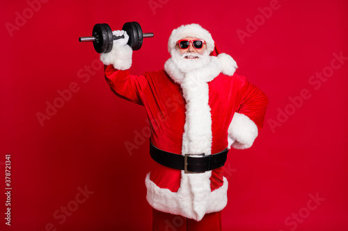 Photo of pensioner old mad man white beard lift heavy dumbbell easily shiny smiling confident self-assured hand hip wear santa x-mas costume sunglass cap isolated red color background
