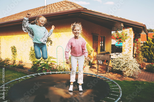 Happy two blonde girls are jumping on a trampoline in the garden near their house. Leisure and games