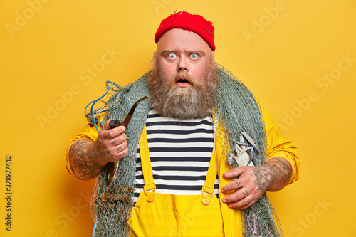 Plump bearded professional man mariner stares at camera with displeased shocked expression holds smoking pipe and goes fishing isolated on yellow background. Fatso seaman stunned to see storm at ocean