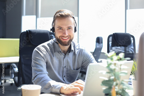 Smiling male call-center operator with headphones sitting at modern office, consulting online information in a laptop, looking up information in a file in order to be of assistance to the client.