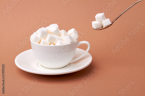 White cup of coffee is overfilled with sugar cubes. Too sweet beverage concept. Isolated on brown surface.