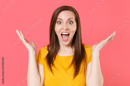 Shocked amazed excited surprised young brunette woman wearing yellow casual t-shirt posing keeping mouth open say wow spreading hands looking camera isolated on pink color background studio portrait.