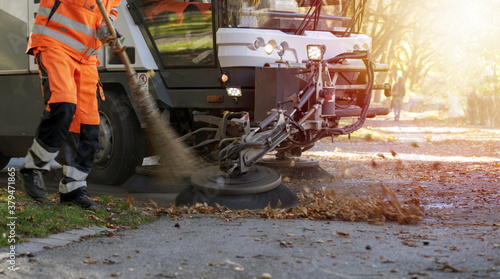 New generation of small electric street sweeper removing fallen leaves in body at autumn city park. Municipal urban services using ecology green vehicle lorry to clean streets from foliage.