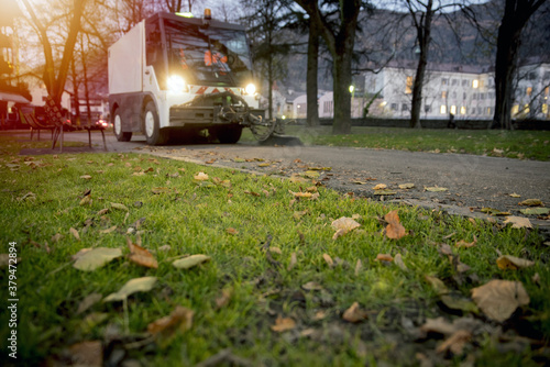 New generation of small electric street sweeper removing fallen leaves in body at autumn city park. Municipal urban services using ecology green vehicle lorry to clean streets from foliage.