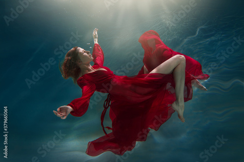 A slender girl in a long red dress swims underwater.