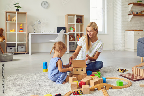 Young mom playing with cute little son and teaching him to build toy house in cozy modern apartment