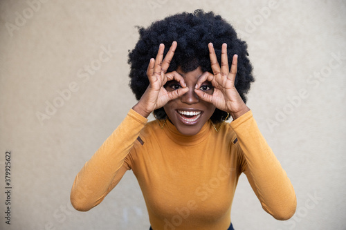 Portrait of cheerful African American woman with curly hair, makes okay gesture over eyes, wearing casual clothing, smiles broadly, completely agrees with suggestion, posing on light background.