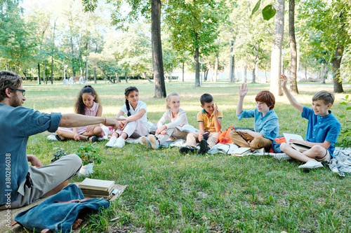 Young teacher in glasses pointing at pupil while listening to his answers during outdoor class