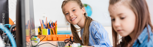 Panoramic shot of schoolgirl looking at camera near friend and computers in stem school