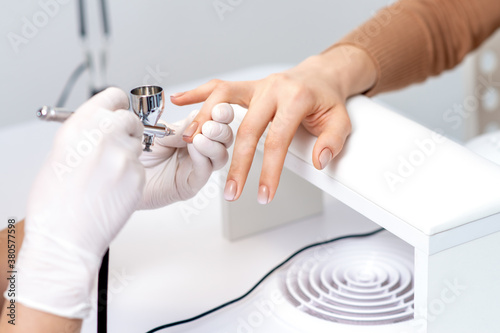 Young woman receiving manicure by airbrush in nail salon. Procedure for spraying paint on the nails