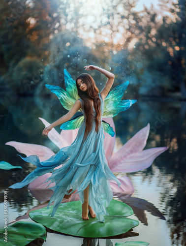 Beautiful young fantasy woman in the image of a river fairy dances on a water lily flower. A long silk dress flies in the wind, butterfly wings glisten. Background evening dark nature, blue lake.