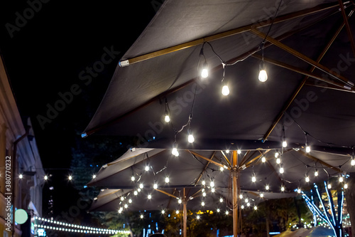textile umbrella with wooden frame and edison string lights glowing with white light on backyard terrace, cityscape night scene with garland, nobody.