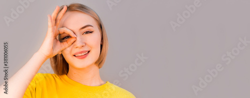 Funny amusing woman in yellow sweater showing okay gesture near her eye