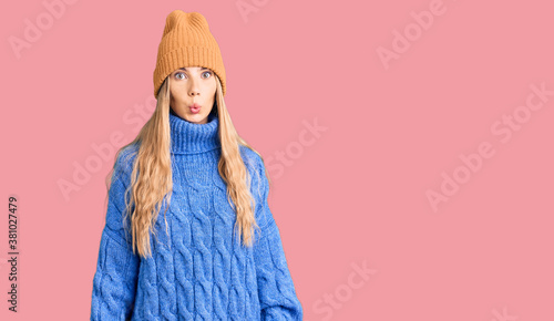 Beautiful caucasian woman with blonde hair wearing wool sweater and winter hat scared and amazed with open mouth for surprise, disbelief face