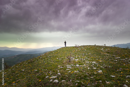 Mountain hiker standing at the edge of the peak coverd by green grass and yellow flowers and photographing distant mountains under a stormy, dark clouds on the sky