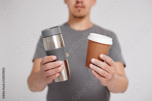 zero waste and eco friendly concept - young man comparing thermo cup with disposable paper coffee cup over grey background