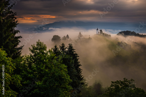 Jamnik, Slovenia - Magical foggy summer sunrise at Jamnik St.Primoz church. The fog gently goes by the small chapel with colorful sky and Julian Alps at background