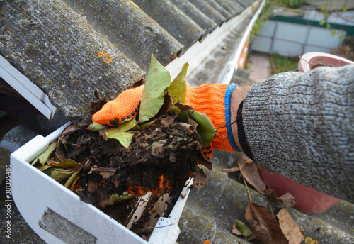 A man is cleaning a clogged roof gutter from dirt, debris and fallen leaves to prevent water damage and let rainwater drain properly.