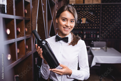 Selective focus of sommelier holding bottle of wine and looking at camera near racks