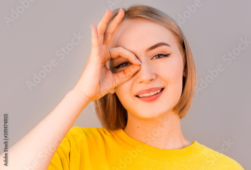 Funny amusing woman in yellow sweater showing okay gesture near her eye