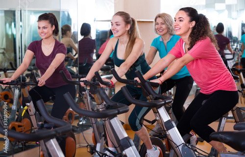 Group of cheerful females of different age training on exercise bikes at fitness club . Selective focus..