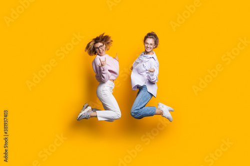 Two blonde sisters jumping on a yellow studio wall and smile at camera gesturing happiness