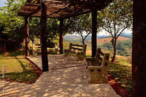 three benches made of cement and wood, under a pergola, in a lookout overlooking a green valley.