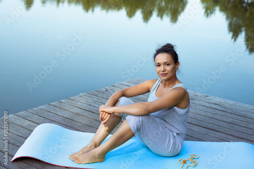 A young woman sits freely on a mat on the shore of the lake and looks at us. Yoga class in nature.