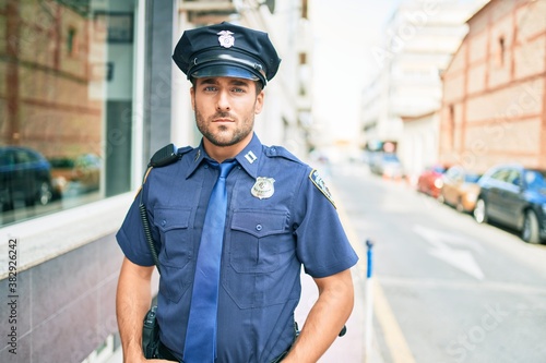 young handsome hispanic policeman wearing police uniform. Standing with serious expression at town street.