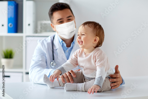 medicine, healtcare, pediatry and people concept - happy doctor or pediatrician wearing face protective mask for protection from virus disease with baby on medical exam at clinic