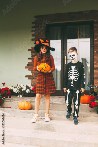 Brother and sister celebrate halloween on the street near the house in costumes and make-up with a pumpkin lantern, play and laugh