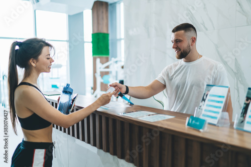 Happy caucasian female client of gym satisfied with service and friendly conversation with employee, slim pretty woman in active wear holding card on reception talking to hospitable receptionist