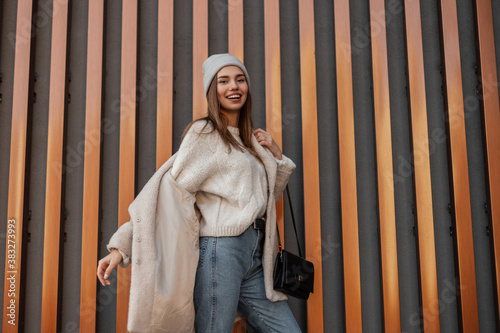 Cheerful young smiling woman with a positive smile in stylish warm clothes with a black leather handbag takes off a fashionable coat walks near a metal gold building. Attractive happy girl model.