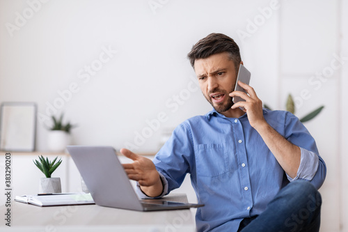 Angry businessman talking on cellphone and looking at laptop screen in office