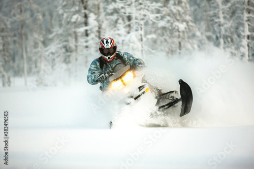 The rider in gear with a helmet makes a sharp turn on a snowmobile on a deep snow surface on a background of snowy landscaping nature and winter forest.