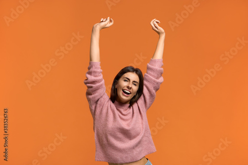 Happy sound.Energized happy woman in pink knitted sweater raises hands joyfully, being in high spirit, jumps playfully, dances to favourite music. Isolated on orange wall