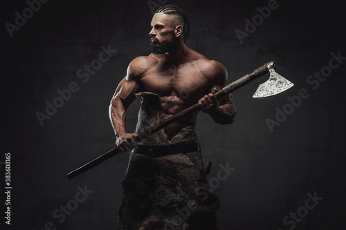 Furious and bearded viking warrior with naked torso and huge biceps prepared to fight holding his weapon in dark background.