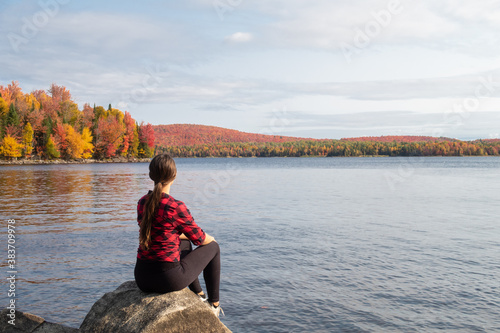 Back view of a young woman sitting on a rock and admiring a lake in the Frontenac national park, Canada