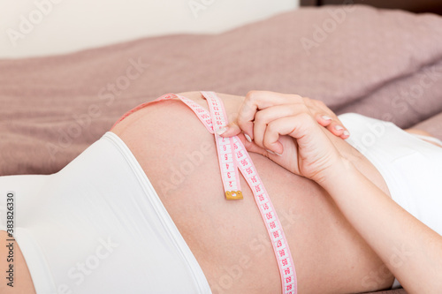 Young beautiful pregnant girl measuring her belly with a tape in home on bed. Healthy pregnancy concept