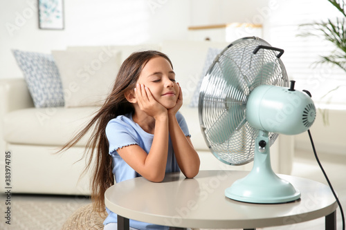 Little girl enjoying air flow from fan at home. Summer heat