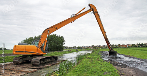 A crane on a pontoon is dredging a canal in an agricultural polder landscape (meadows). 