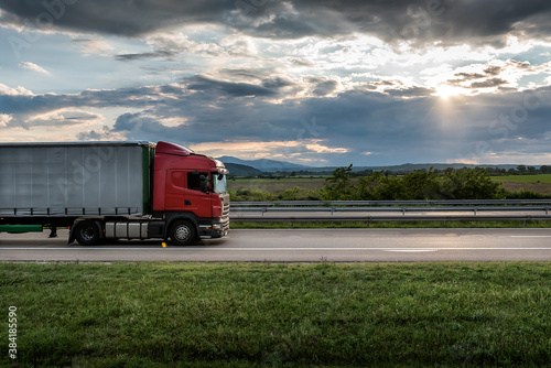 Red truck is on highway - business, commercial, cargo transportation concept, beautiful sunset sky, clear and blank space - side view