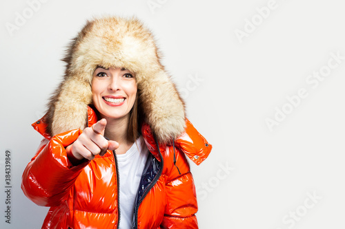 happy young girl in a red jacket and a fur hat, looks at the camera and points a finger in front of him on a light background. Concept gestures, advertising, fashion