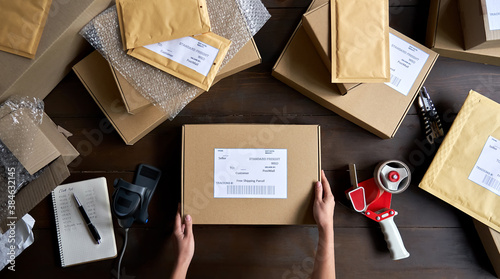 Above table top view of female warehouse worker or seller packing ecommerce shipping order box for dispatching, preparing post courier delivery package, dropshipping shipment service concept.