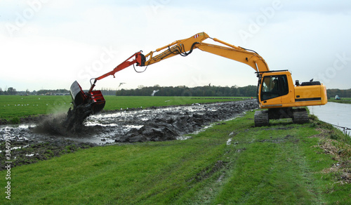 Dredging a canal by a crane with clamshell bukcet in a Dutch polder landscape; dredging spoil is deposited on land.