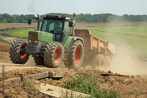 A tractor with empty tipper trailer drives up a dike on a dusty track; in the background a green polder and a reenforced dike with a new layer of clay. 