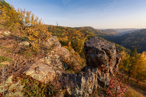 Autumn landscape in golden colors. In the foreground there are rocks, trees with yellow leaves. In the background, mountains covered with forest in the autumn haze.