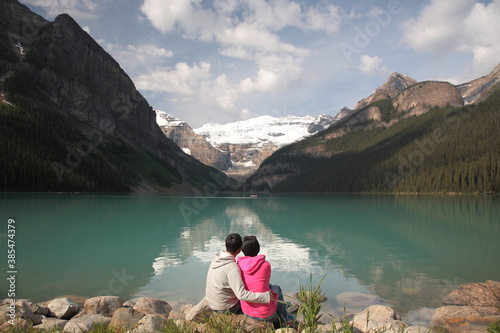 Couple siting on rock by the lake watching Mount Victoria with Victoria Glacier and Lake Louise during summer in Banff National Park, Canadian Rockies, Alberta, Canada.
