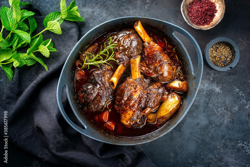 Modern style traditional braised slow cooked lamb shank in red wine sauce with shallots and carrots offered as top view in a design stewpot